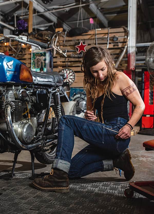 A woman with long dreadlocks sits in a garage, working on a vintage motorcycle. The setting is rustic with various tools and motorcycle parts around. She wears jeans, a black tank top, and brown boots, focused on her task. Image courtesy of Nathalie Dupré Photography.